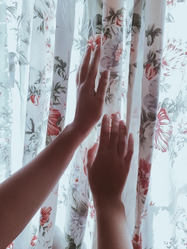 A persons hand touching a white curtain with floral pattern.