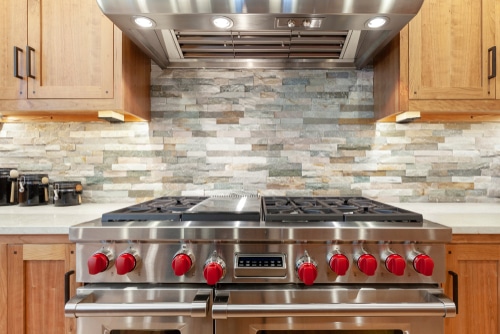 A mosaic of brick stones against a large stainless oven with red knobs.